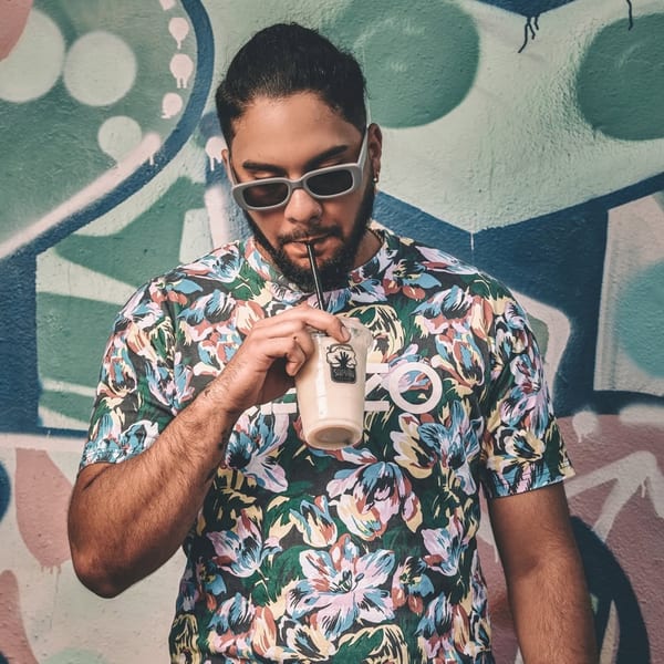Man in sunglasses and floral shirt sips a Saman drink against a colorful graffiti wall.