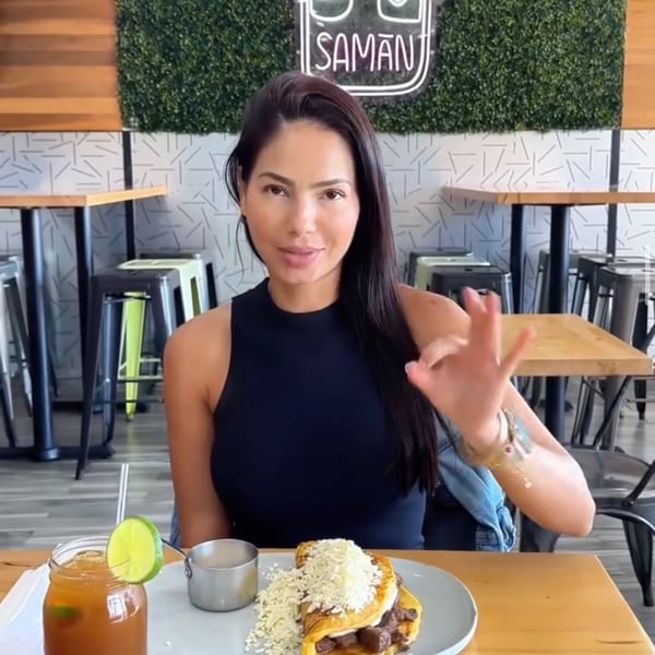 Woman at Saman restaurant with a Venezuelan cachapa, drink, and making an "OK" sign.
