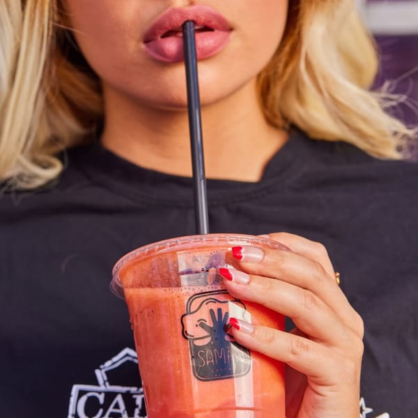 Blonde woman with red-tipped nails sips a red Saman smoothie through a black straw.