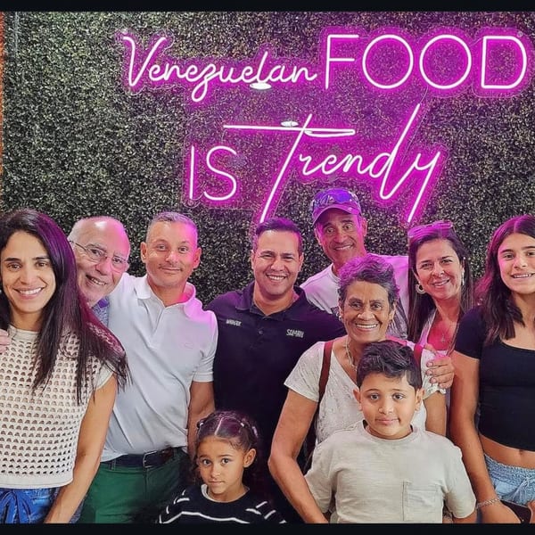 A large group of people, including children, smiling in front of a neon sign that reads "Venezuelan FOOD IS Trendy".