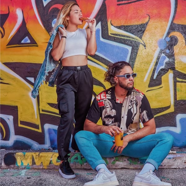 A woman eats a churro while a man sits beside her, both in front of a graffiti wall.