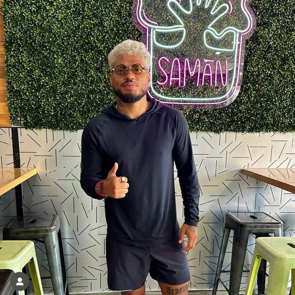 Man with blonde hair and round glasses giving a thumbs up in front of a Saman neon sign.