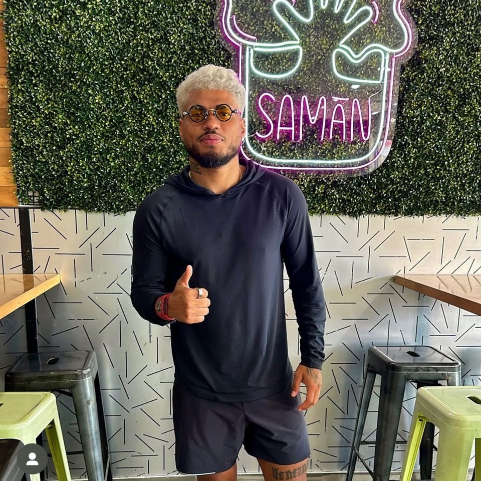 Man with blonde hair and round glasses giving a thumbs up in front of a Saman neon sign.