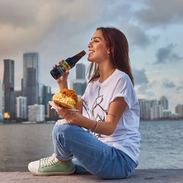 Woman enjoying a Venezuelan arepa and drink by the water with a city skyline.