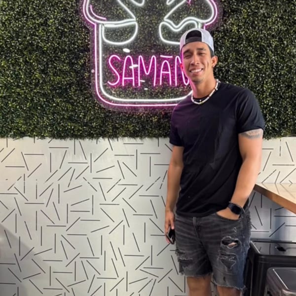 Smiling man in a cap and black shirt standing in front of a neon Saman sign.