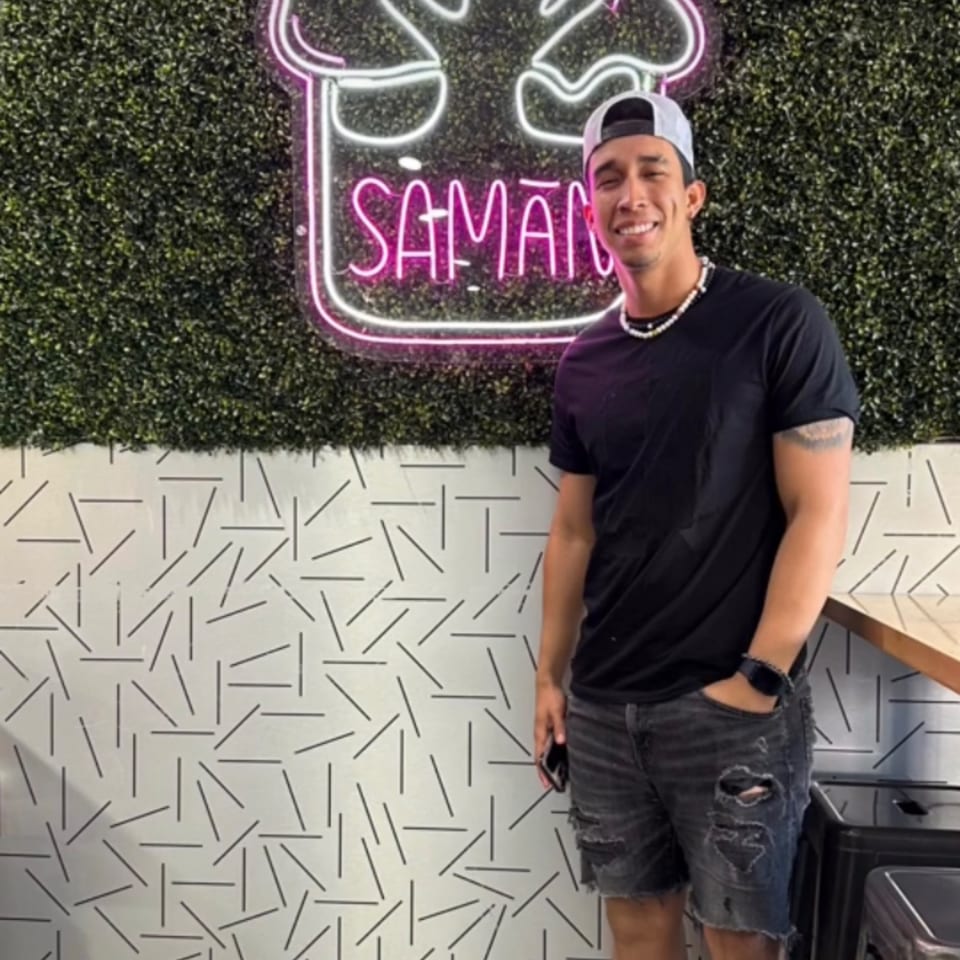 Smiling man in a cap and black shirt standing in front of a neon Saman sign.