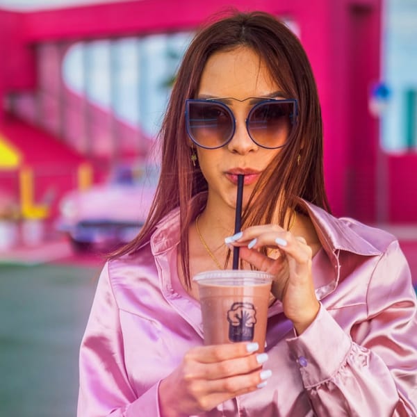 Woman in sunglasses and a pink satin shirt sips a Saman drink through a straw.