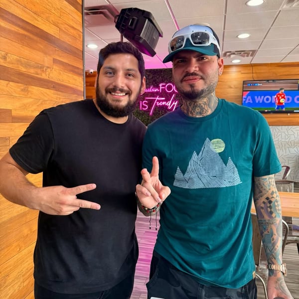 Two men smiling and making peace signs, with a neon sign and TV in the background.